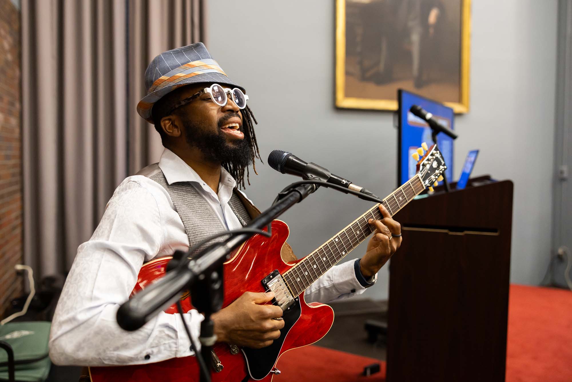 Photo: A Black man sitting, playing guitar, singing in a mic. He wears a blue hat and red pants.