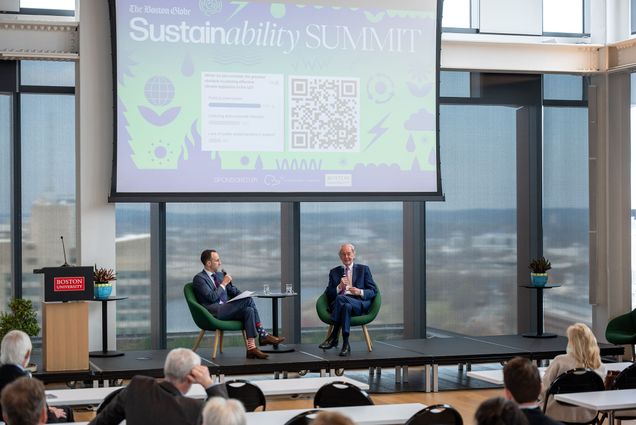 Photo: Two men sit on a stage with a projector above them with a screen that reads SUSTAINABILITY SUMMIT