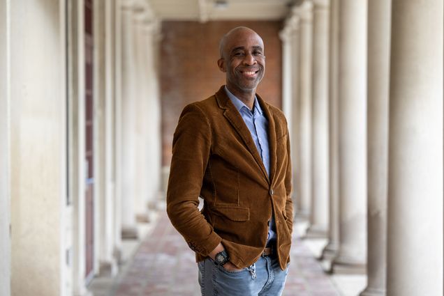 Photo: A man wearing a corduroy jacket poses for a formal portrait outside on a sunny day, in a row of columns