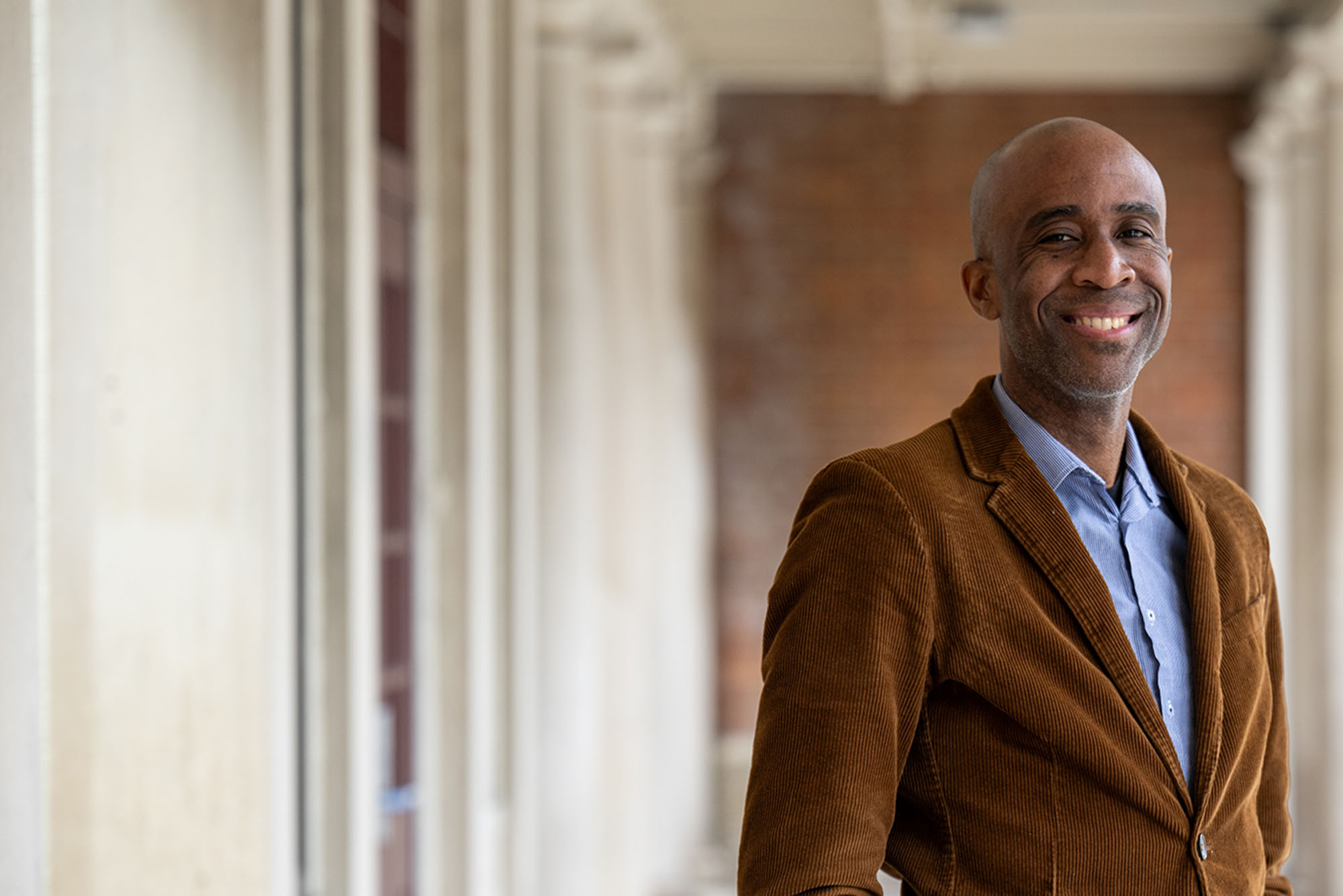 Photo: A man wearing a corduroy jacket poses for a formal portrait outside on a sunny day, in a row of columns