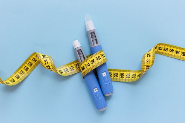 Photo: Two injectors for anti-obesity medication with yellow measuring tape wrapped around them in front of a blue background