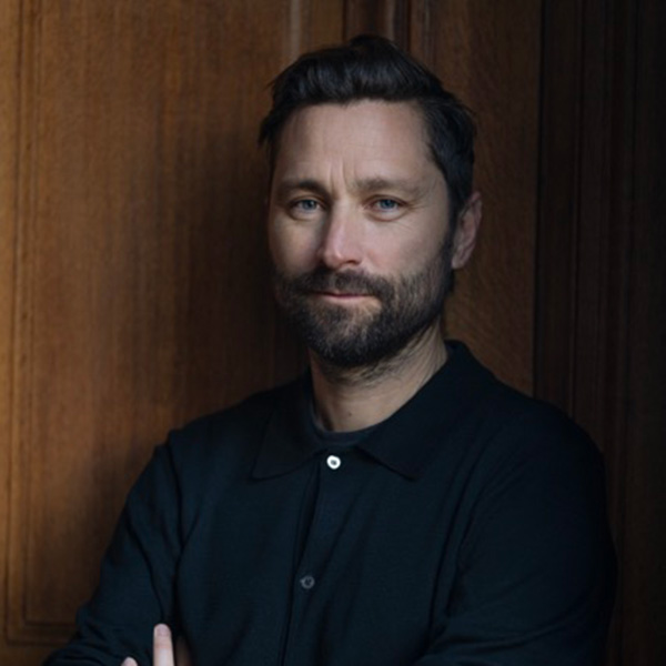 Photo: A man stands with arms crossed in a black shirt for a formal portrait