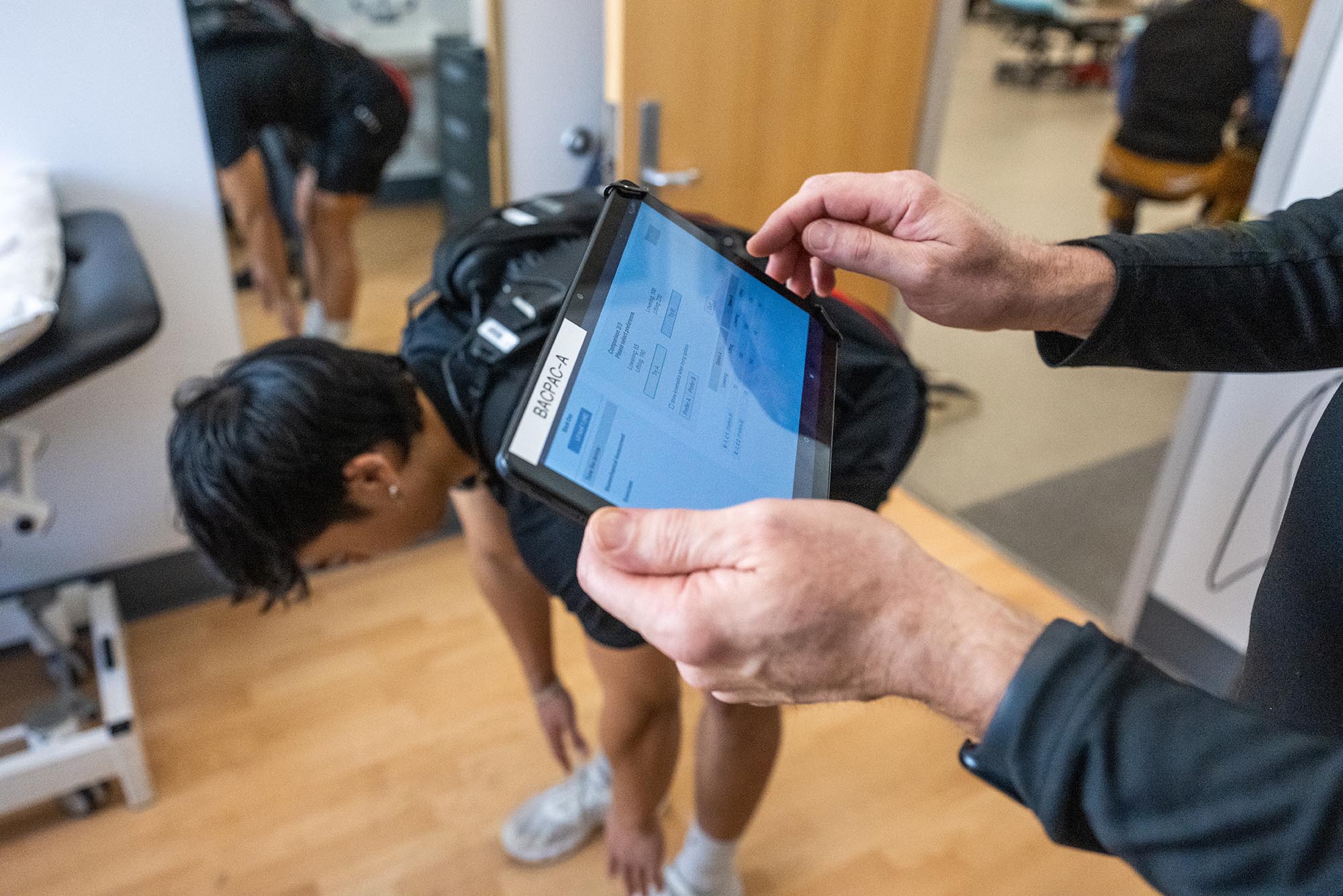 Photo: A person observes an iPad with metrics on the screen while a person behind the iPad stretches towards the ground.