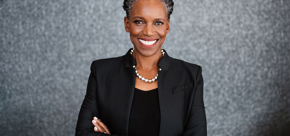 Photo: A Black woman with a short grey and black afro and wearing a necklace of large, faux metal pearls, black blouse, and black blazer, smiles and poses with arms crossed in front of a grey backdrop.