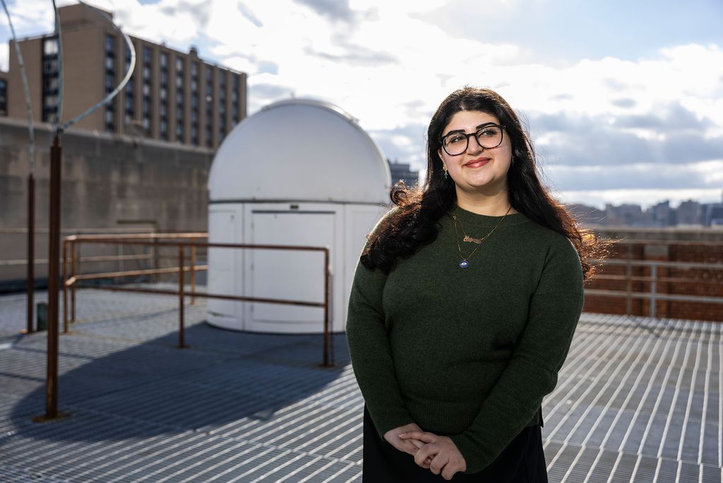 Photo: A female student standing on a rooftop with a closed smile, looking off in the distance