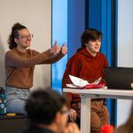 Photo: Two people sitting in front of a larger group, one on the left is giving a thumbs up and the other sits in front of a computer