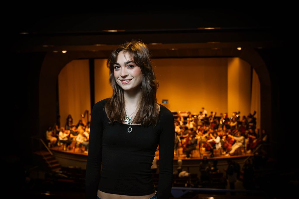 Photo: A girl with brown hair and bangs wearing a long sleeve black shirt and smiling in front of a stage