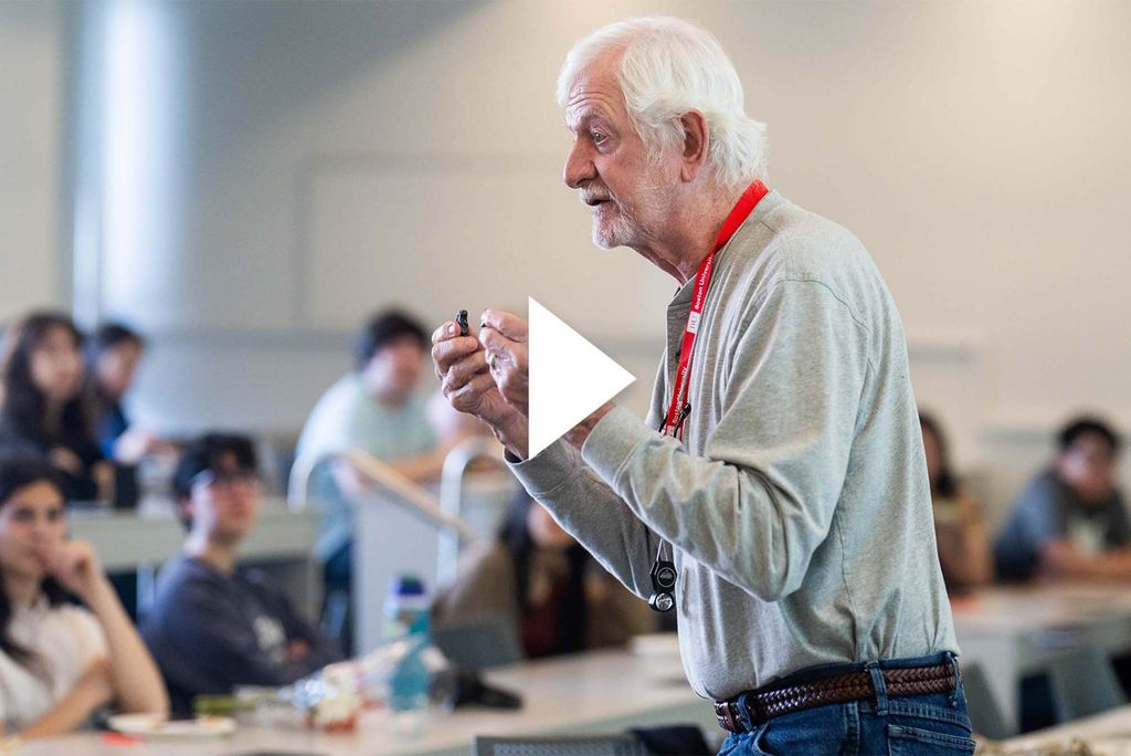 Photo: An older man in jeans and a buttoned shirt holds a rock as he teaches a class.
