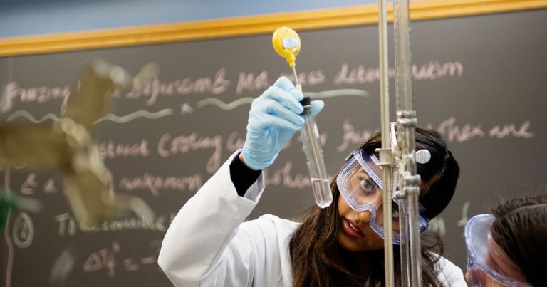 A student wearing goggles examines a test tube.