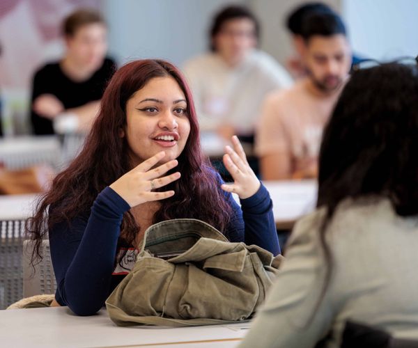 Woman sits across the table from another student speaking and gesturing with her hands