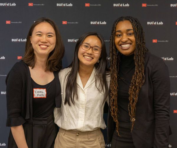 Three women smiling together standing in front of a BU backdrop