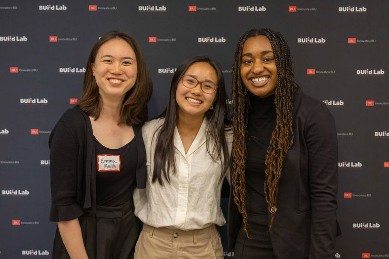 Three women smiling together standing in front of a BU backdrop