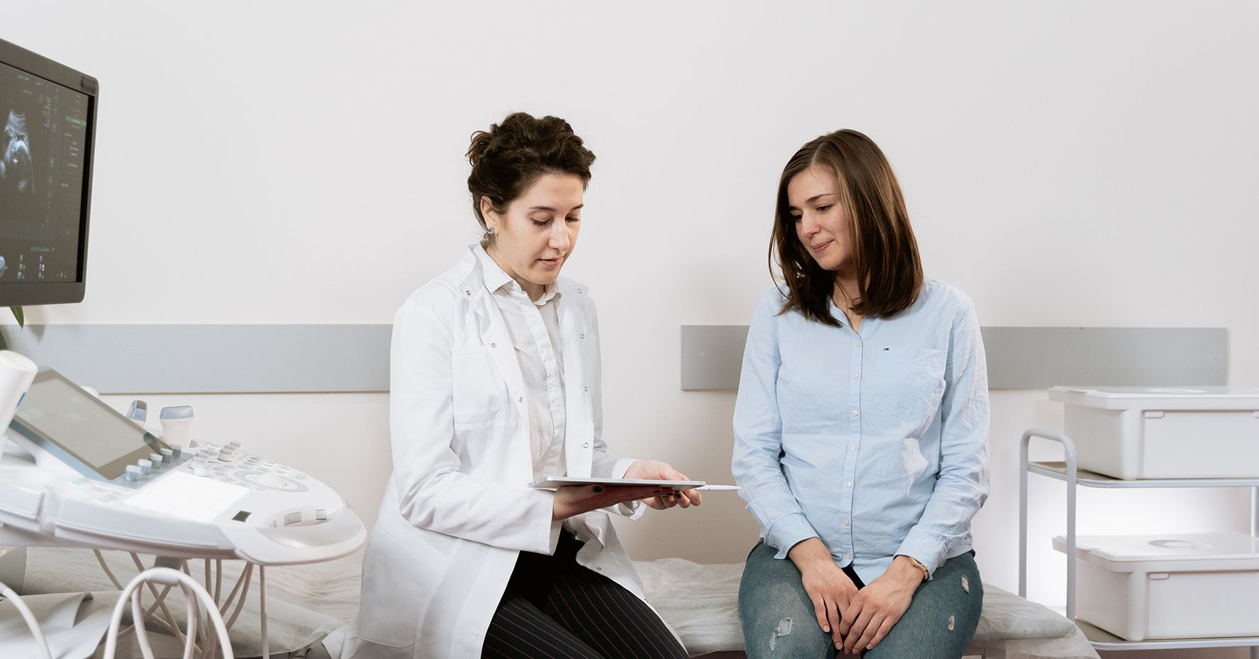 A female doctor in a lab coat talks with a female patient in an exam room.