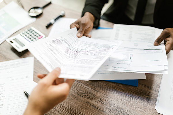 Tax law at BU law. Two people exchange tax forms on a desk. The hands, calculator, and magnifying glass appear on the desk.