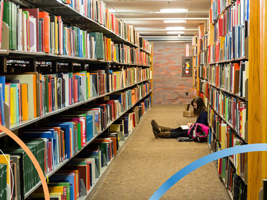 A Boston University student working on a laptop on the floor between library shelves