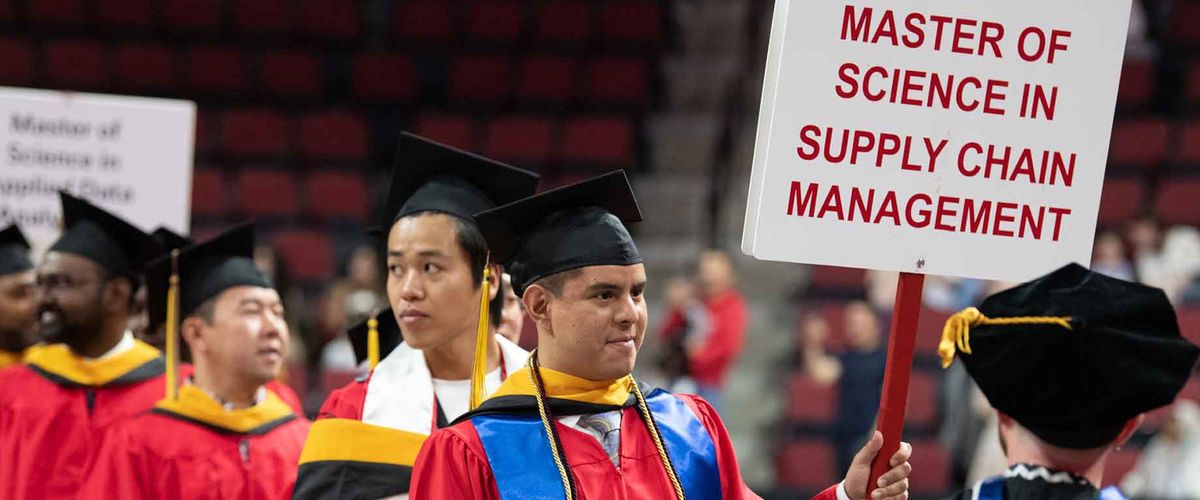 A graduate walking during the MET convocation ceremony, holding a sign that reads “Master of Science in Supply Chain Management”