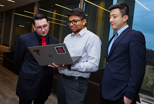 Three standing students huddling around a laptop