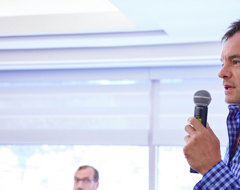 Man leaning against wall at front of room speaking with microphone