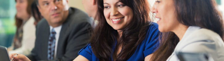 Mid adult Hispanic woman is studying latop computer screen during a job training business conference. She is reviewing financial information with coworker as they sit in a row with other professional businesspeople. Women are pointing at computer screen and smiling.