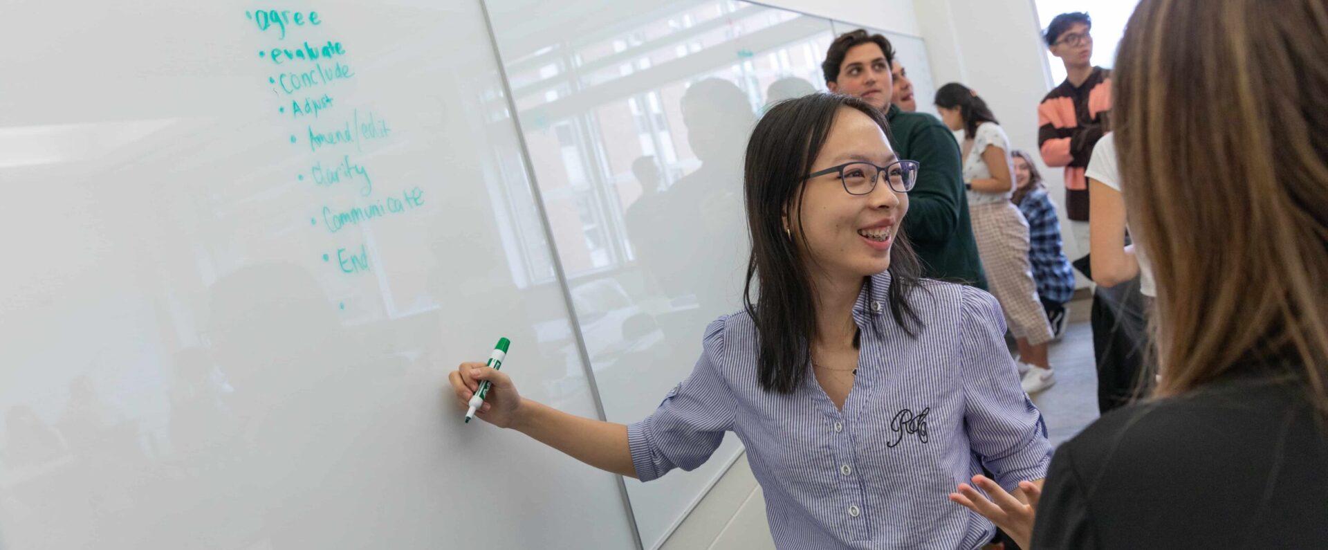 Three smiling students sit in class and work together. The one in focus wears red. 