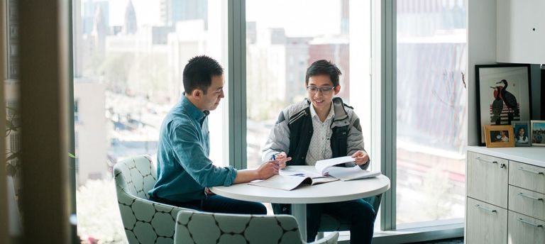 Two people sit at a table in front of a window. They are looking at papers together and having a lively discussion.