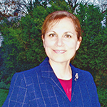 A woman with Brown hair stands in front of green foliage. She is wearing a blue checked blazer with a gold brooch with a pink shirt.