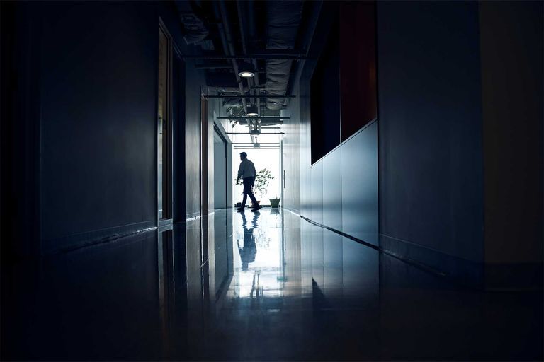 A person walks down a hallway. They are backlit by a bright window, but the hallway is otherwise dark.