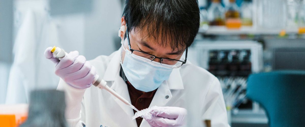 A person with short dark hair wearing a white lab coat, glasses, a mask and gloves performs pipette work in a lab.