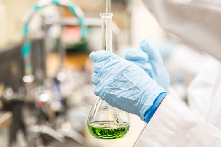 A person wearing gloves and a lab coat holds a beaker and pipette