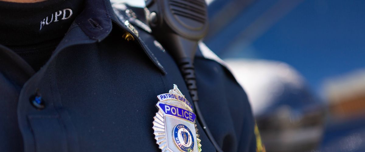 The image shows a close-up of a police officer's uniform. Prominently displayed is a badge that reads "Patrol Officer, Police, Boston University, Comm. of Massachusetts." The officer is also wearing a dark shirt with a microphone clipped to the shoulder area, and the initials "BUPD" can be seen on the turtleneck collar.
