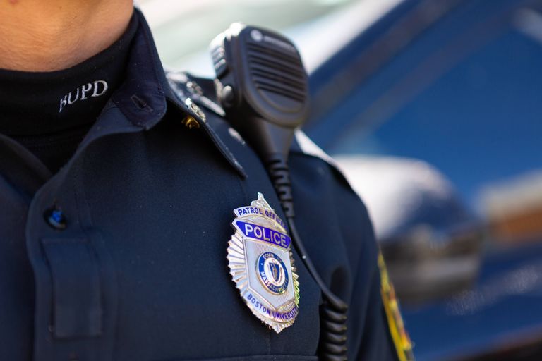 The image shows a close-up of a police officer's uniform. Prominently displayed is a badge that reads "Patrol Officer, Police, Boston University, Comm. of Massachusetts." The officer is also wearing a dark shirt with a microphone clipped to the shoulder area, and the initials "BUPD" can be seen on the turtleneck collar.