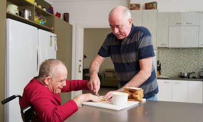 Senior man standing in kitchen, buttering bread for another senior man in a wheelchair.