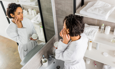 african american woman applying cosmetic cream on face and looking in mirror in bathroom
