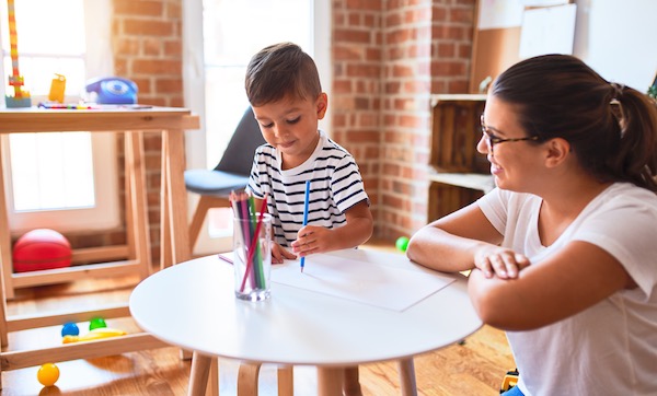 Beautiful teacher and toddler boy drawing draw using colored pencils at kindergarten