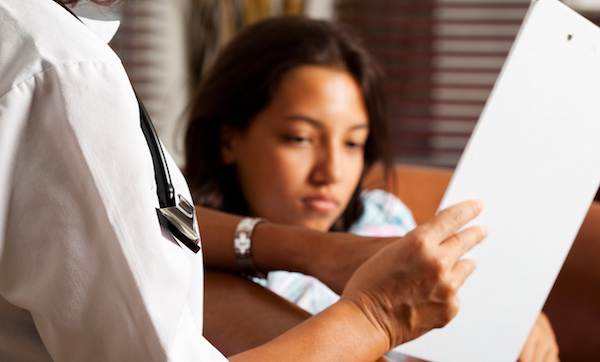 Doctor holding a clipboard, showing it to her child patient and explaining something.