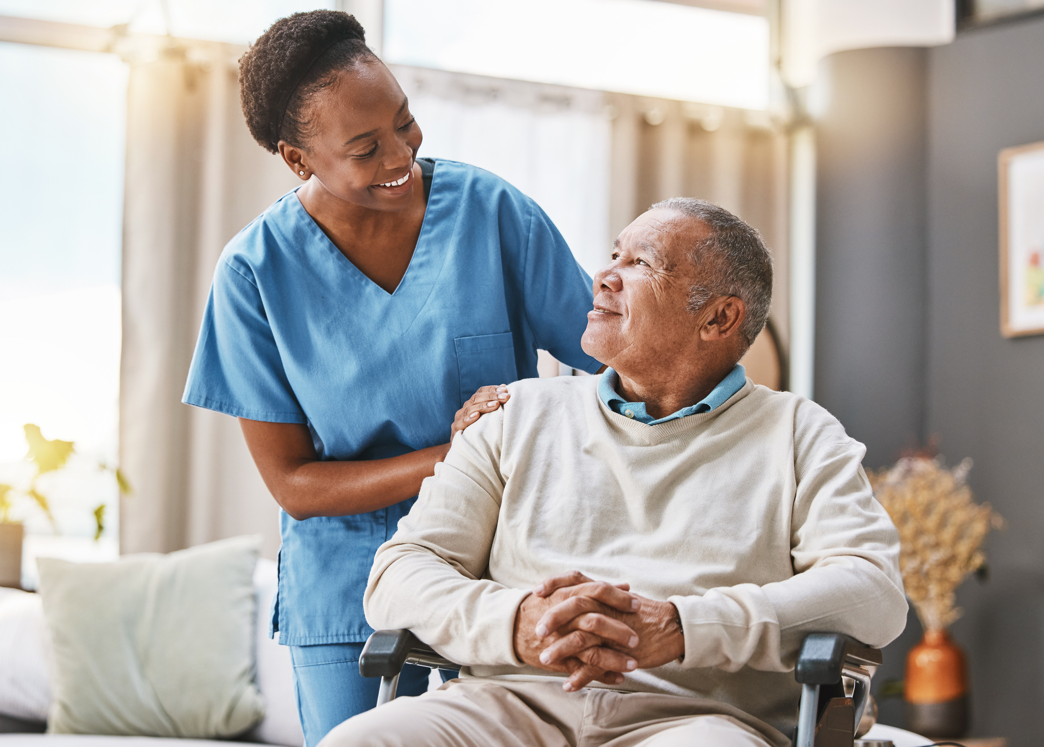 Elderly man sits in a chair and smiles up at nurse with her hand on his shoulder