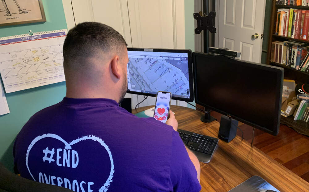Stephen Murray, wearing a shirt that says #end overdose, looks at his phone while sitting at his desk