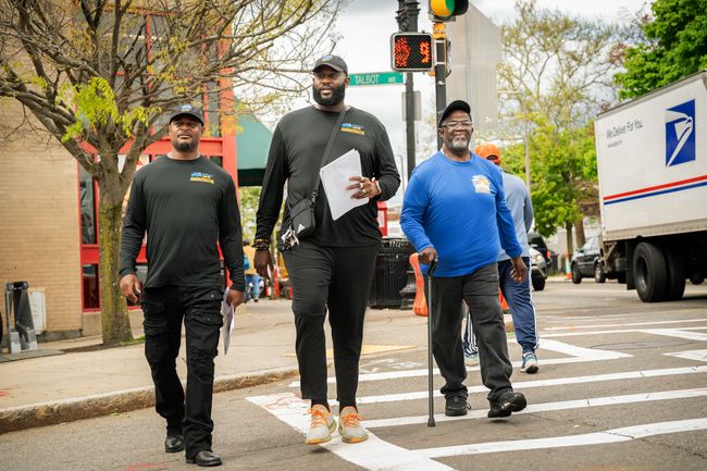 3 Black men cross the street wearing t-shirts with "Fathers Uplift" on them.