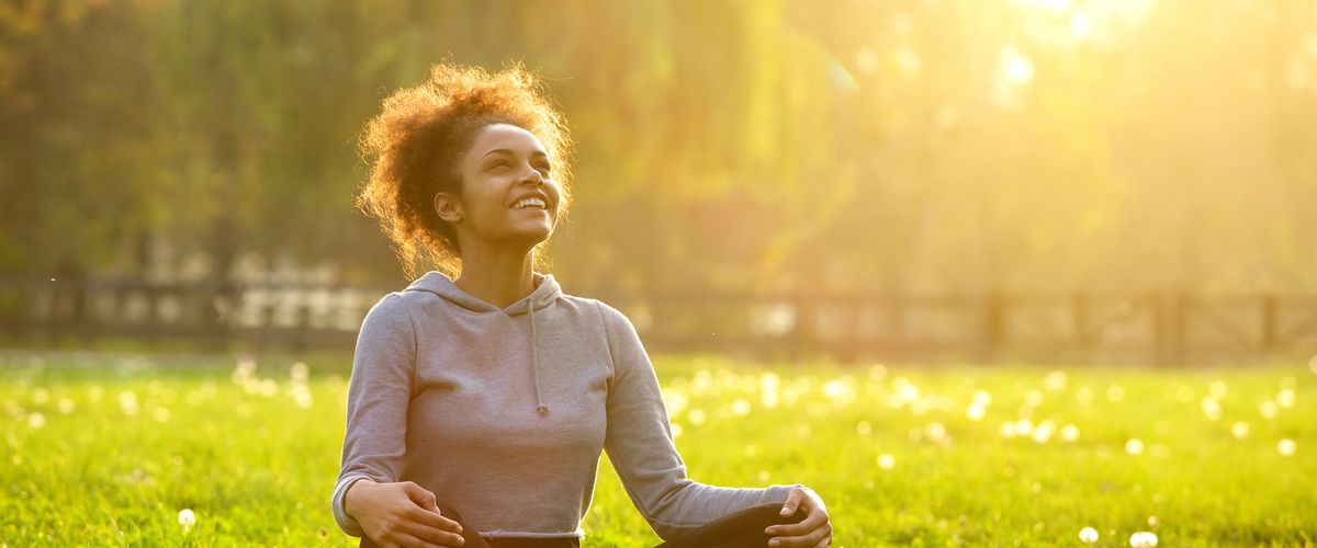 Woman sitting crosslegged in a sunny field while smiling and looking up to the sky.