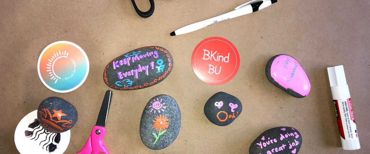 several stones decorated with happy images and wellness messages sit on a table with stickers, scissors, and pens.