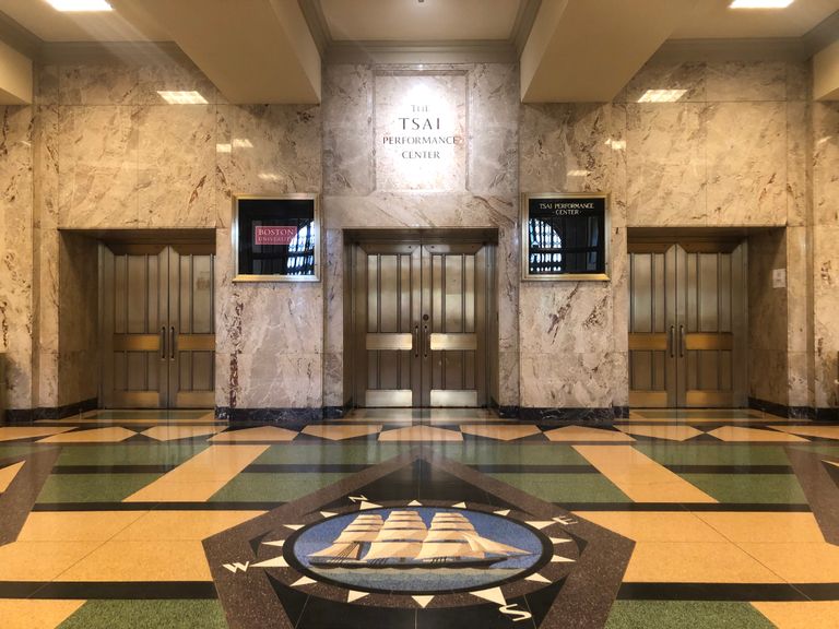 Lobby with tall ship image inlayed in marble floor in front of 3 sets of brass doors with word "Tsai Performance Center" etched over center doors.
