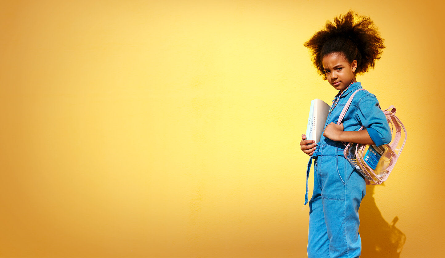 Side view of stylish African American girl with books and school bag looking at camera while standing against yellow background during studies