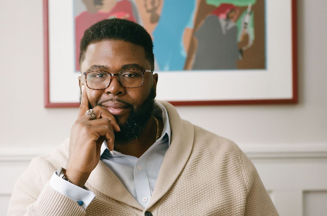 Shows Anthony Jack wearing a white collared shirt and beige sweater, he has short hair and is wearing glasses