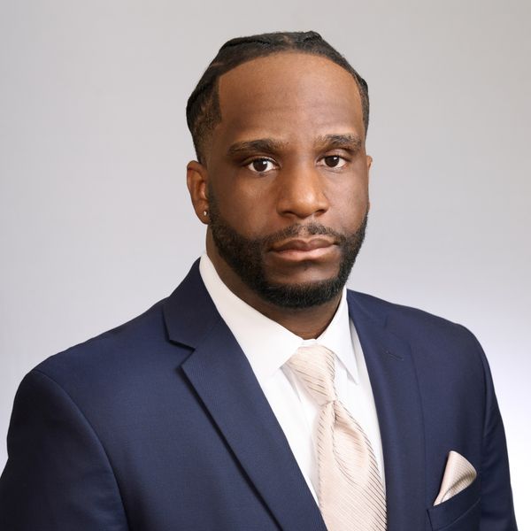 Shows Joram Dade, who has short black hair and beard, and is wearing a navy jacket, white collared shirt, and cream colored tie