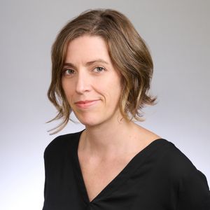 Shows Naomi Caselli, who has short light brown hair and is earing a v neck black top