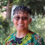 Shows Gloria Thomas, who has short curly hair and is wearing a brightly colored top and glasses