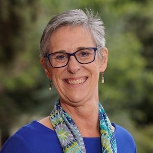 Shows Sherri Hughes, who has short gray hair and is wearing blue rimmed glasses, a blue shirt, and a multicolored scarf