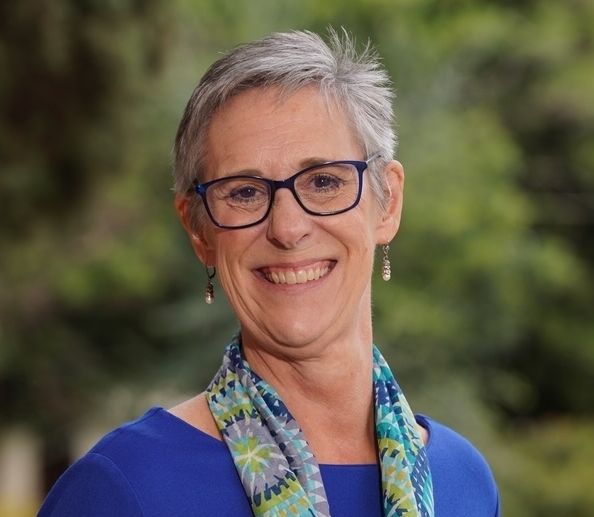 Shows Sherri Hughes, who has short gray hair and is wearing blue rimmed glasses, a blue shirt, and a multicolored scarf