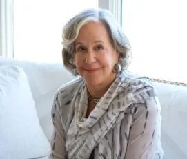 Kathryn Lasky sitting on a cream couch smiling wearing a grey sweater