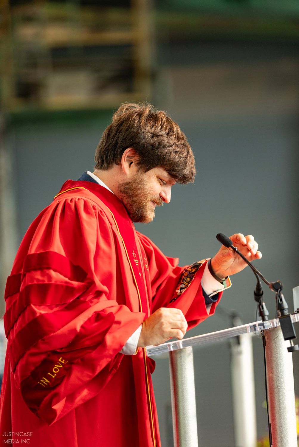 A photo of Adam Bush speaking in a red doctoral robe